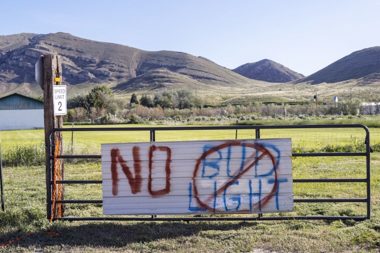 A board reading "No bud light" is posted on a fence overlooking a rural field