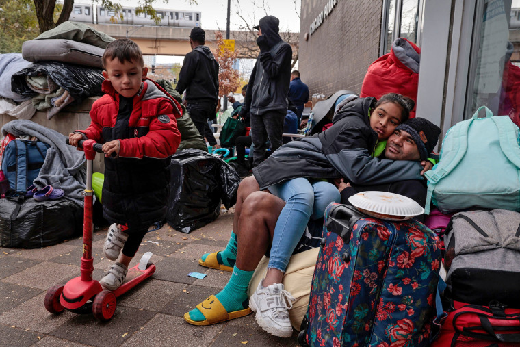 A migrant child scooters past Venezuelan migrants Michael Castejon and daughter, Andrea Sevilla, outside the 1st District police station in Chicago, on Nov. 3, 2023.