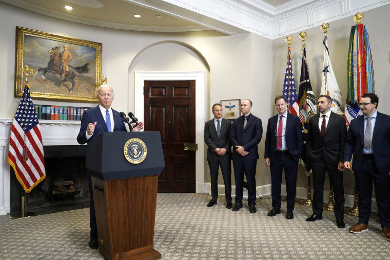 President Joe Biden speaks at a podium while several men in suits look on.