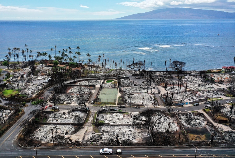 An aerial view of a recovery vehicle driving past burned structures and cars in Lahaina