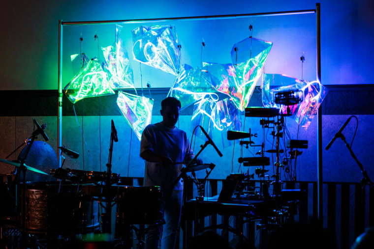 Efraín Rozas, artista musical, está rodeado por instrumentos de percusión y viento frente a una pantalla con formas trigonométricas en colores neón durante una presentación en el Lincoln Center de Nueva York, el 2 de noviembre de 2023