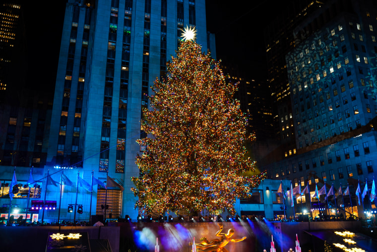 A giant christmas tree stands above a gold statue and ice skating rink surrounded by tall new york city buildings.