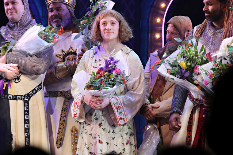 Ethan Slater holds flowers in an old-fashioned floral tunic costume and a bowl-cut wig. He's surrounded by other men in medieval looking knight's outfits also holding flowers.