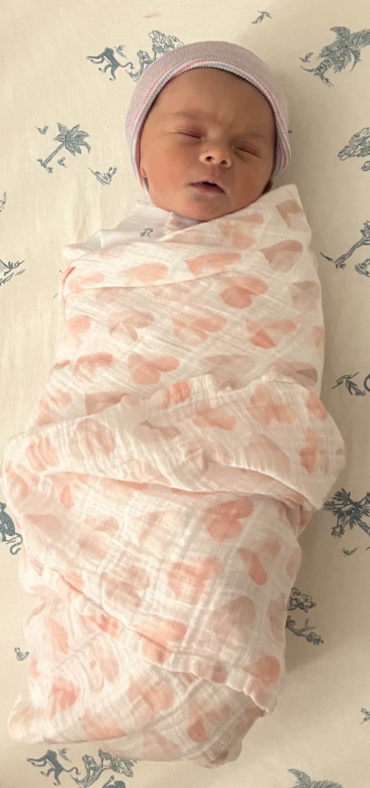 A baby on white and blue sheets swaddled in a white blanket with pink hearts.