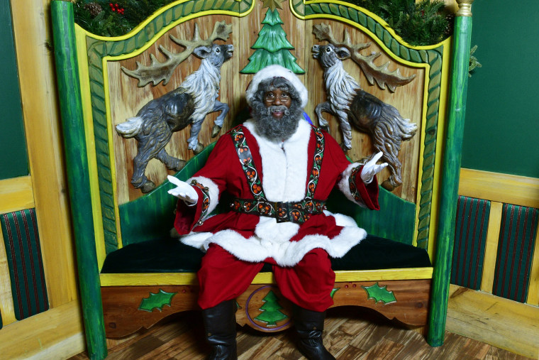 An actor sits dressed in a Santa costume sits in a large wooden chair.