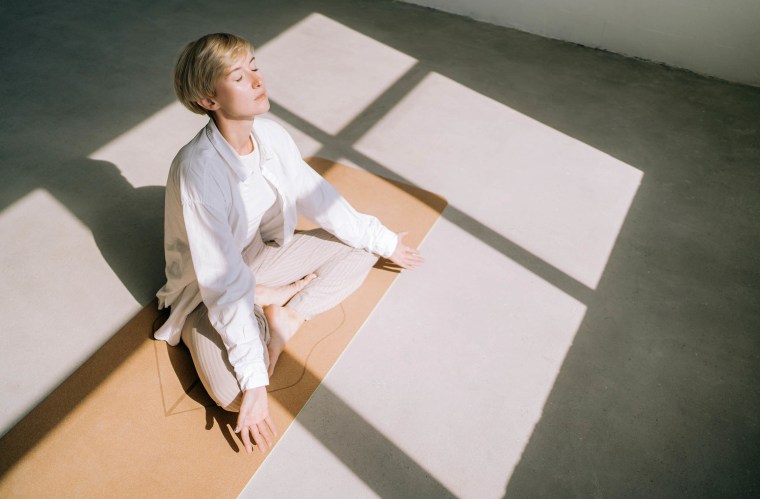 woman with short blond hair is meditating sitting in lotus position on yoga mat in front of a window. 