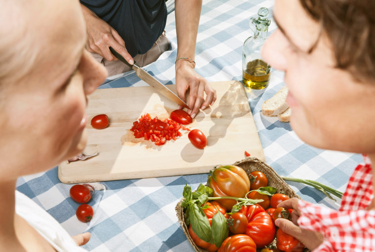 Person cutting tomatoes on table with friends in foreground. 