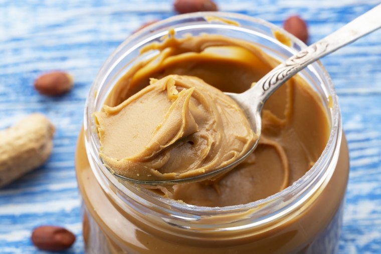 Peanut butter in an open jar and peanuts in the skin are scattered on the blue table.