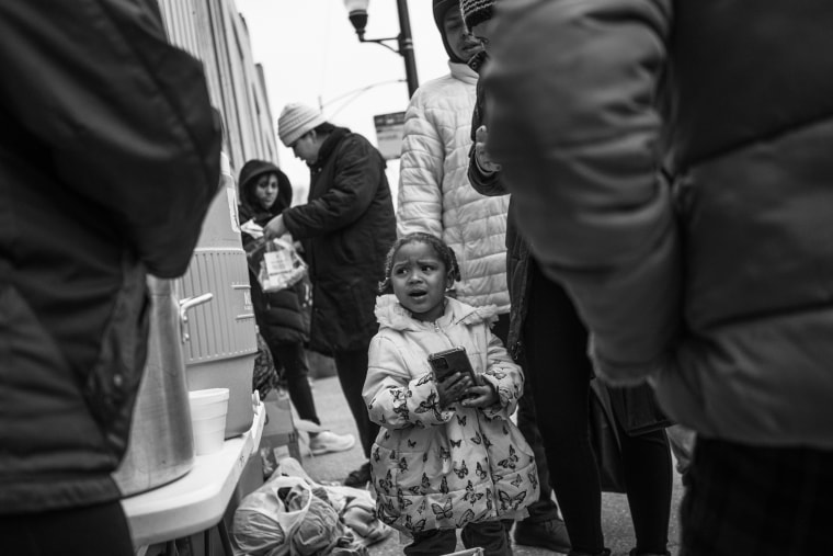 A little girl holds a cell phone as migrants wait in line for oatmeal.