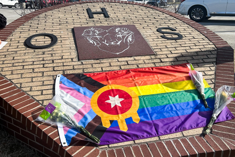 A pride flag and flower bouquets are laid atop the school entry plaque for Owasso High School