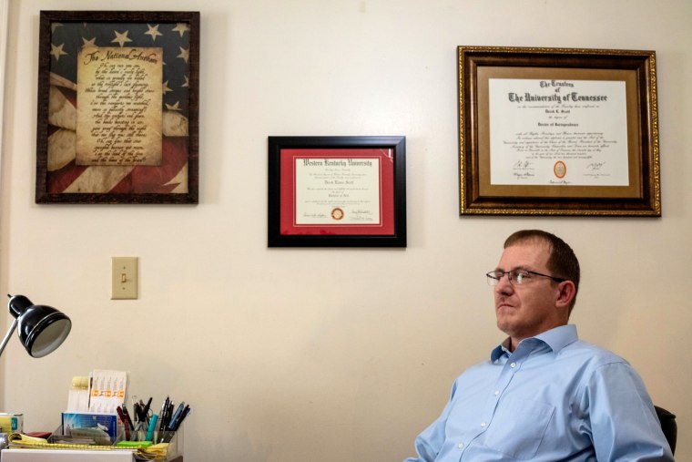 Image: Derek Scott sits at a desk with framed degrees on the wall above him.