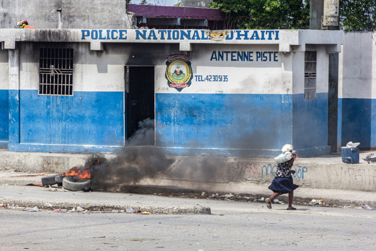 An elderly woman runs in front of the damaged police station building with tires burning in front of it
