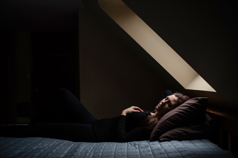 Woman laying on bed with eyes closed in dark room under a sky light.