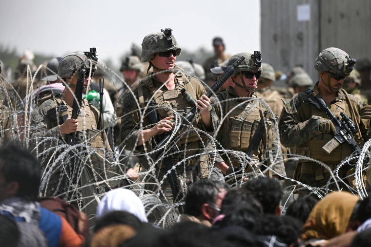 U.S. soldiers stand guard behind barbed wire as Afghans sit on a roadside