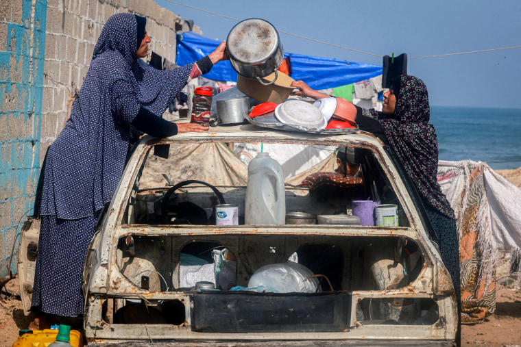 Palestinian women arrange kitchen utensils to dry atop a destroyed car at a makeshift camp for displaced people.