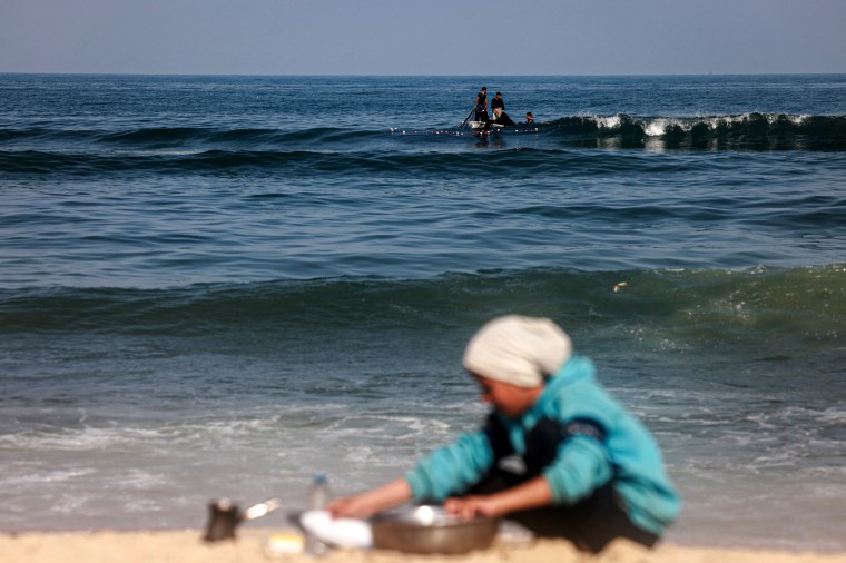 A displaced Palestinian girl washes kitchen utensils on the beach as fishermen sail in a boat.