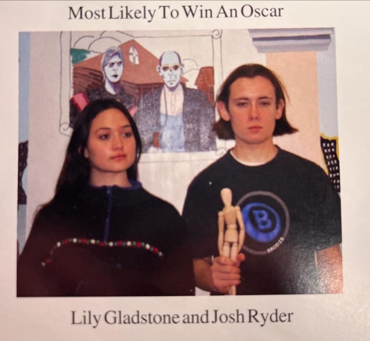A young Lily Gladstone and Josh Snyder hold a wooden statue in front of a mural.