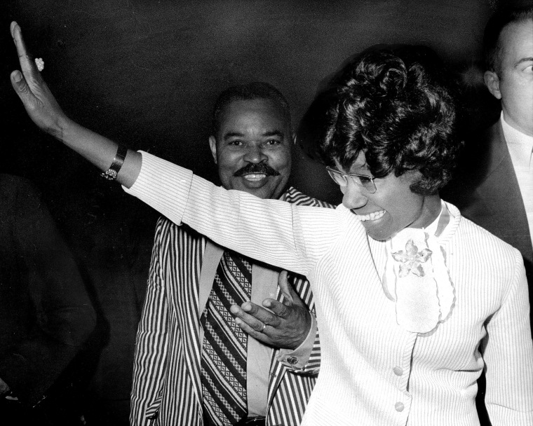Shirley Chisholm waves as her husband stands behind her in a black and white photo.