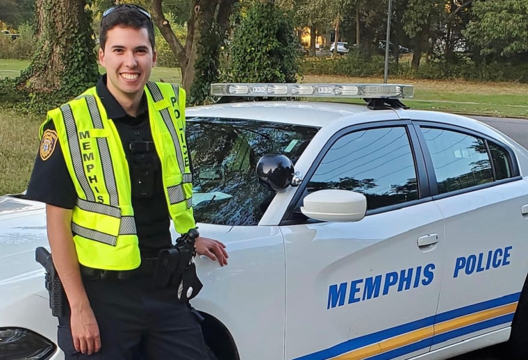Officer Joseph McKinney smiles next to a police car.