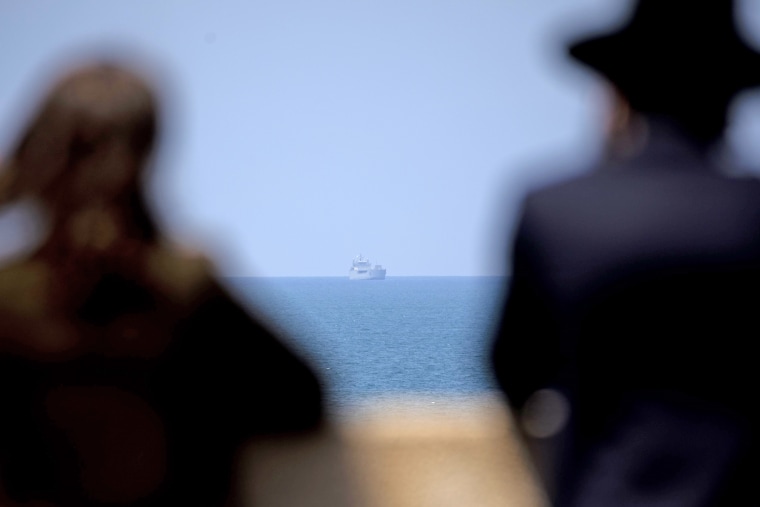 People look at an Israeli naval ship patrolling off the Israeli coast on April 14, 2024 in Natanya.