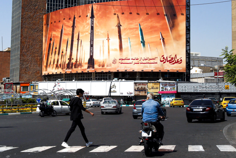 A man crosses a street as motorists drive past a billboard depicting Iranian ballistic missiles in service.