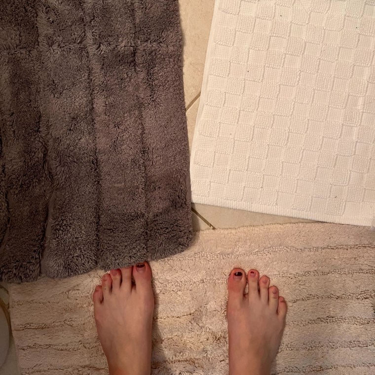 Person stands with feet showing on fluffy beige bath mat with a fluffy brown and checkered white one in front of her.