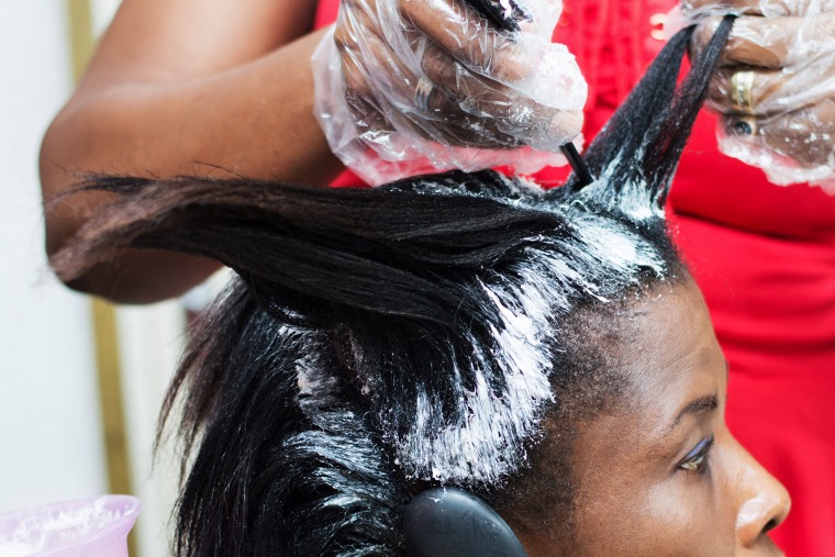 A hairdresser applies hair relaxer to a black woman's roots