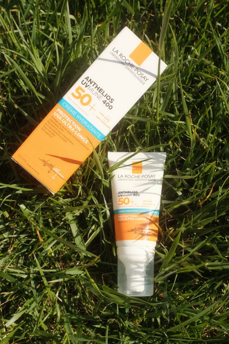 A bottle of sunscreen and its box lay in grass in the sun.