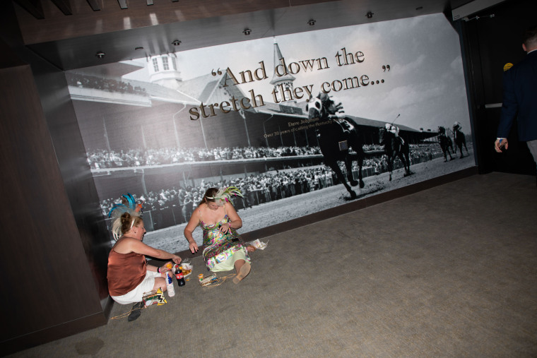 Two women eat near a sign at Churchill Downs that reads "And down the stretch they come..."