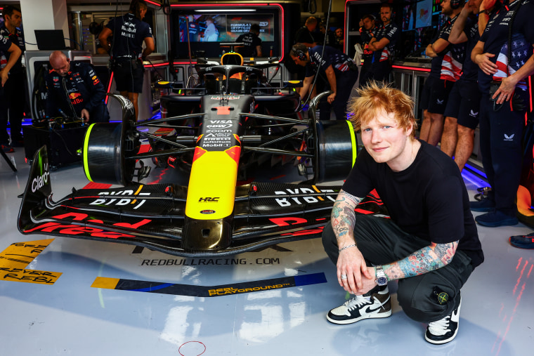 Ed Sheeran crouches next to a race car to take a photo outside the Oracle Red Bull Racing garage