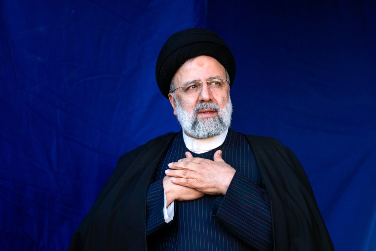 Ebrahim Raisi stands against a blue backdrop and places his hands on his heart