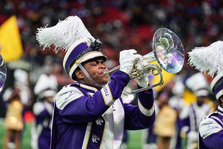 A person wearing a purple and white marching band costume plays the trumpet