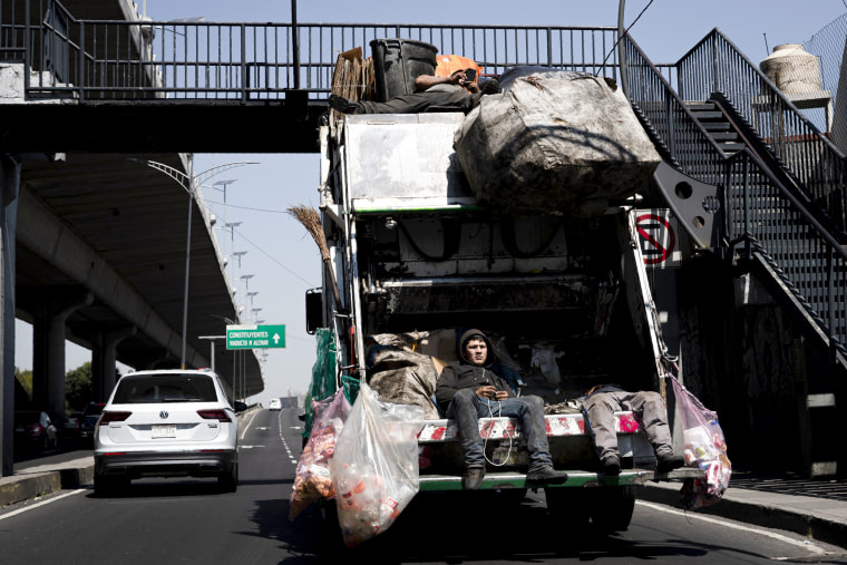 Trash collectors ride on their garbage truck.