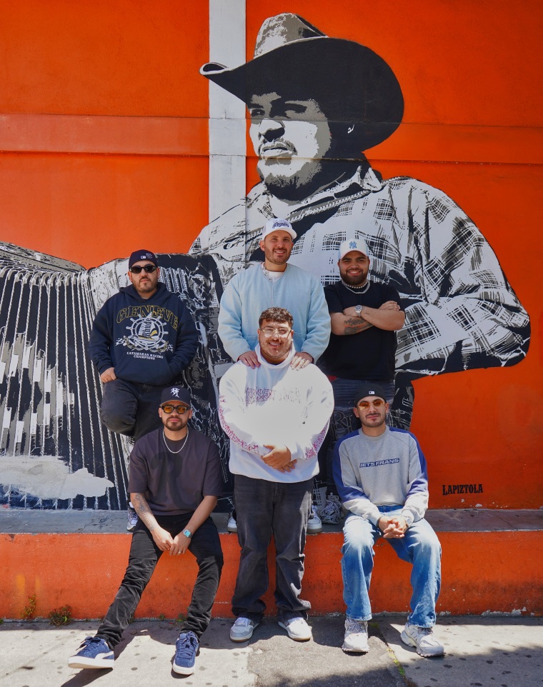 Grupo Frontera pose for a portrait against an orange mural featuring a man in a cowboy hat