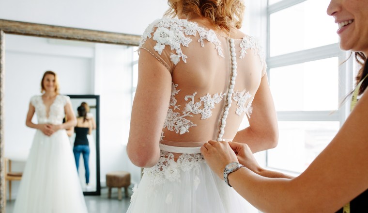 Bride is standing in front of mirror and fitting dress with wedding assistant.