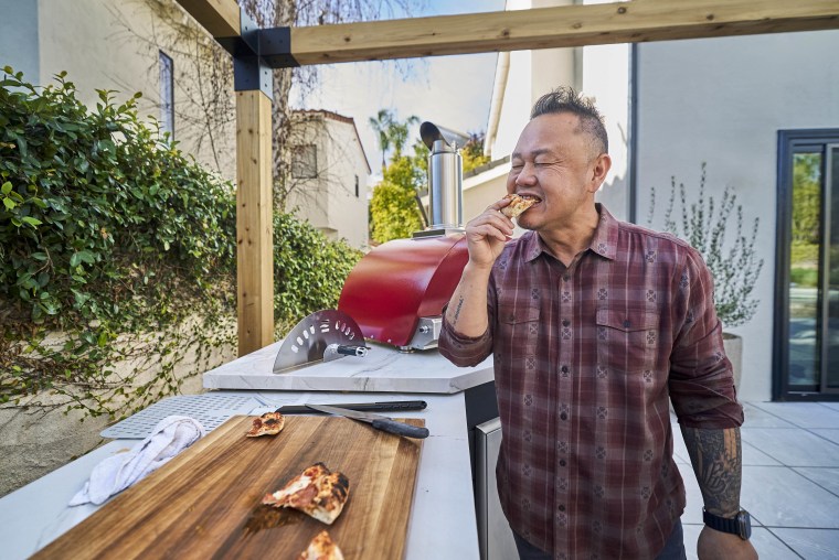 Jet Tila in a flannel shirt bites into a pizza in a backyard outdoor kitchen.