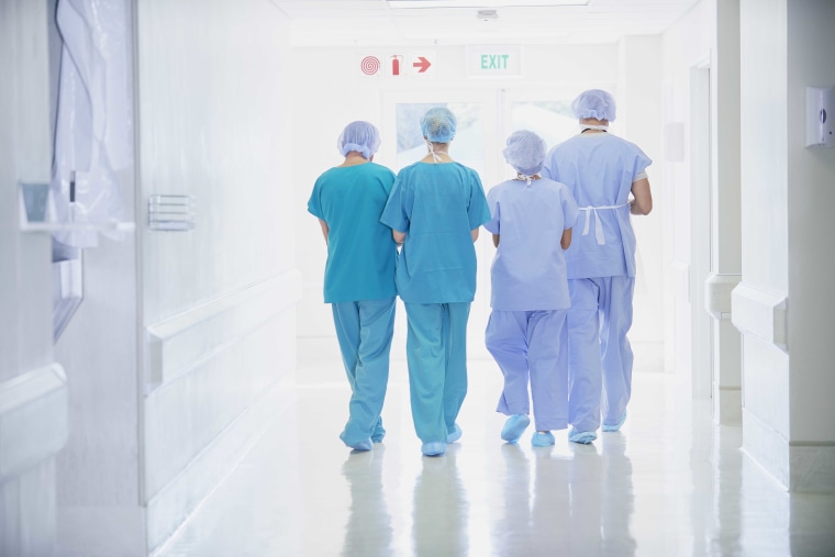 Rear view of four medical staff wearing scrubs walking in hospital corridor.