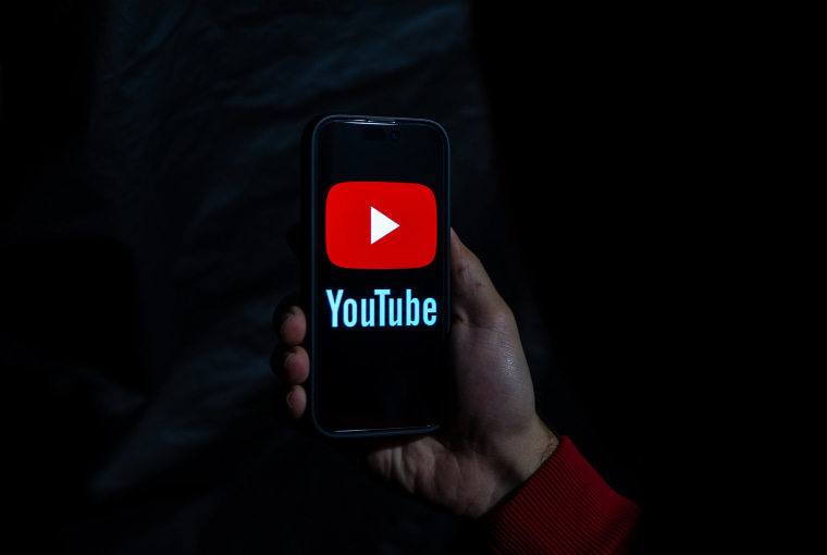 A hand holding a cell phone with the YouTube logo displayed on the screen