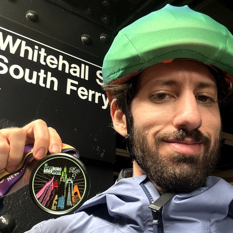 A close up of the author holding a Five Boro Bike Tour medal in front of a subway station sign that reads “Whitehall St-South Ferry”