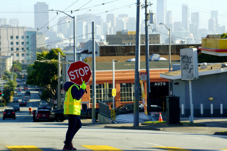 A crossing guard in a yellow vest holds a stop sign in front of their face as they cross the street. The San Francisco skyline is in the background.
