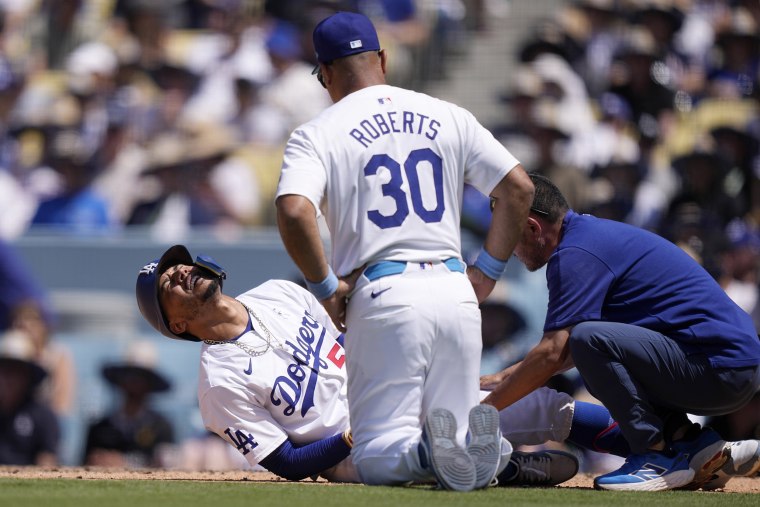 Mookie Betts, left, writhes on the ground after being hit by a pitch as manager Dave Roberts and a trainer tend to him during the seventh inning of a baseball game against the Kansas City Royals, in Los Angeles
