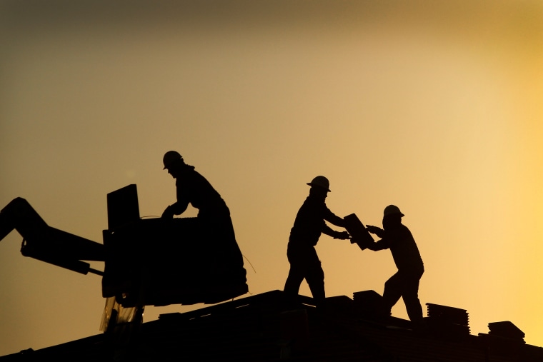 Construction workers pass a bundle of shingles on the roof of a home