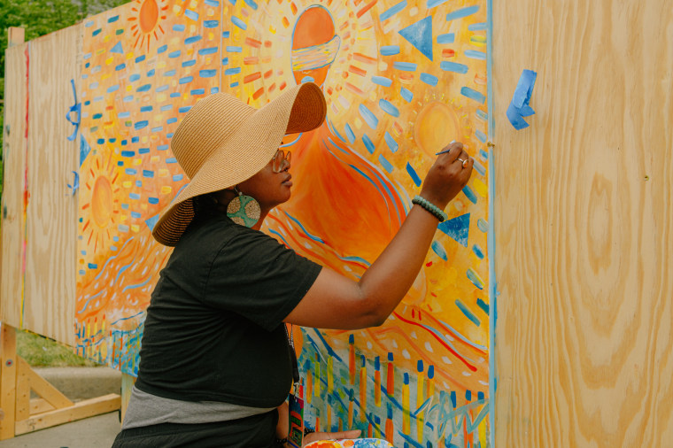 A person wearing a large sunhat paints a mural on a wooden board