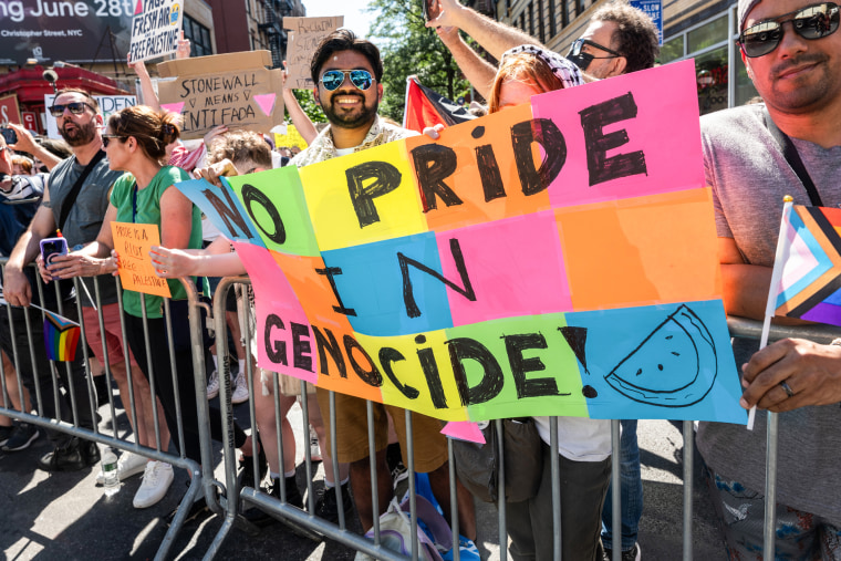 Behind a gate outside, protestors hold a sign that says "No Pride In Genocide"