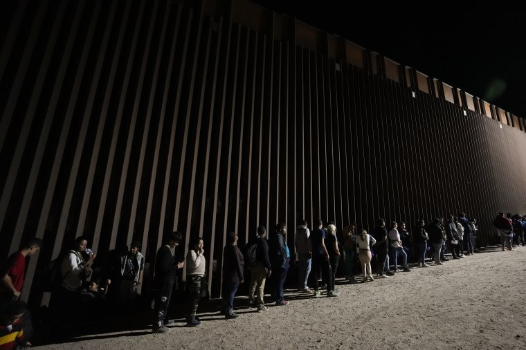 Personas se alinean contra un muro fronterizo mientras esperan para solicitar asilo después de cruzar la frontera desde México, 11 de julio de 2023, cerca de Yuma, Arizona. 