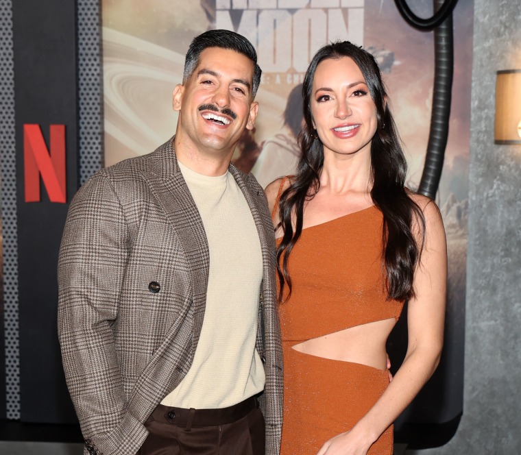 Alexandra Madison and Jon Bouffard pose on a red carpet. Bouffard in a brown plaid jacket and Madison in a rust orange dress.