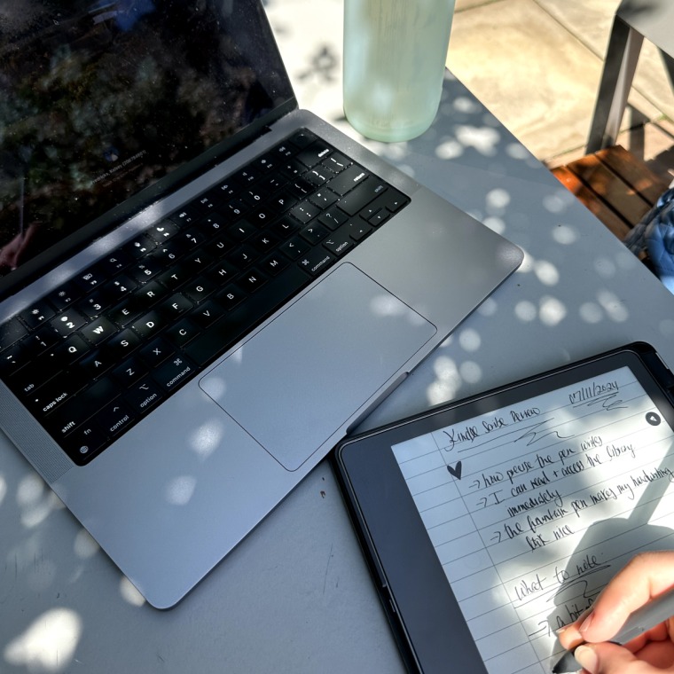 Image of a Macbook and a Kindle Scribe (on an outdoors table in sunny lighting) with some writing on it detailing favorite things and things to note about the Scribe. A water bottle is pictured in the background.