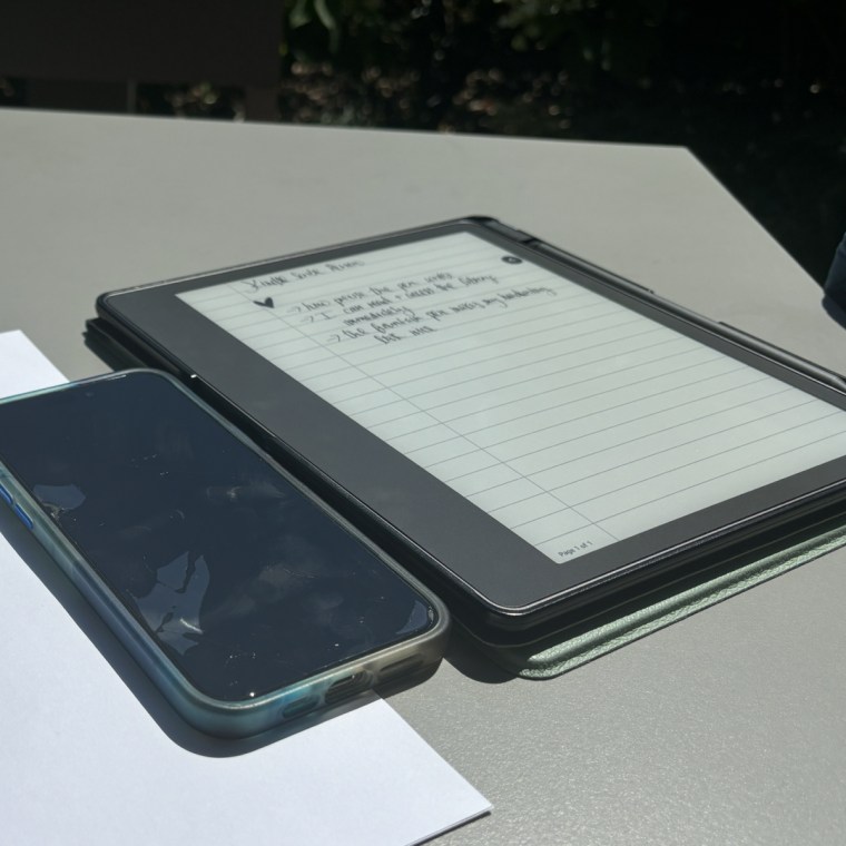 An image of an iPhone and a Kindle Scribe next to each other on a grey table in sunny lighting.