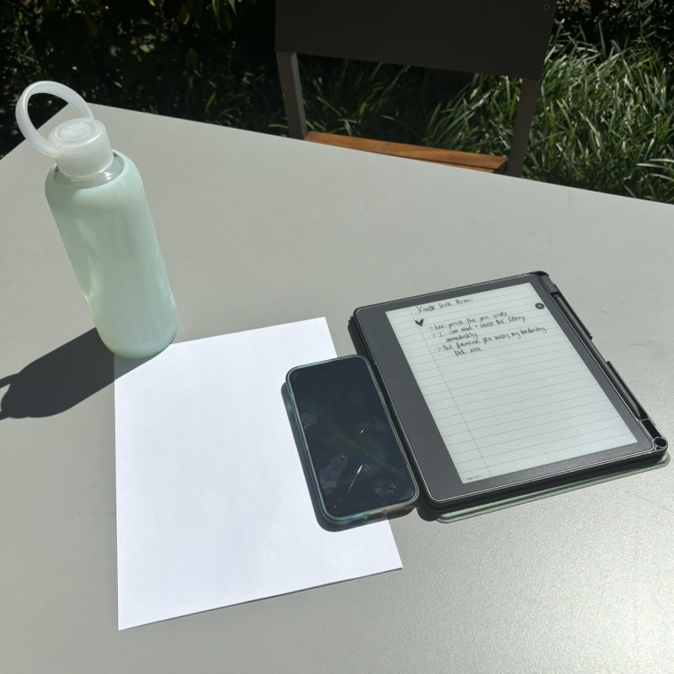 A light teal BKR water bottle, 8.5x11" sheet of paper, an iPhone 14 Pro Max next to the Kindle Scribe on a grey table in bright sunlight.