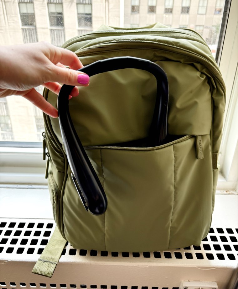 Hand placing a black neck fan into the outside pocket of a green backpack